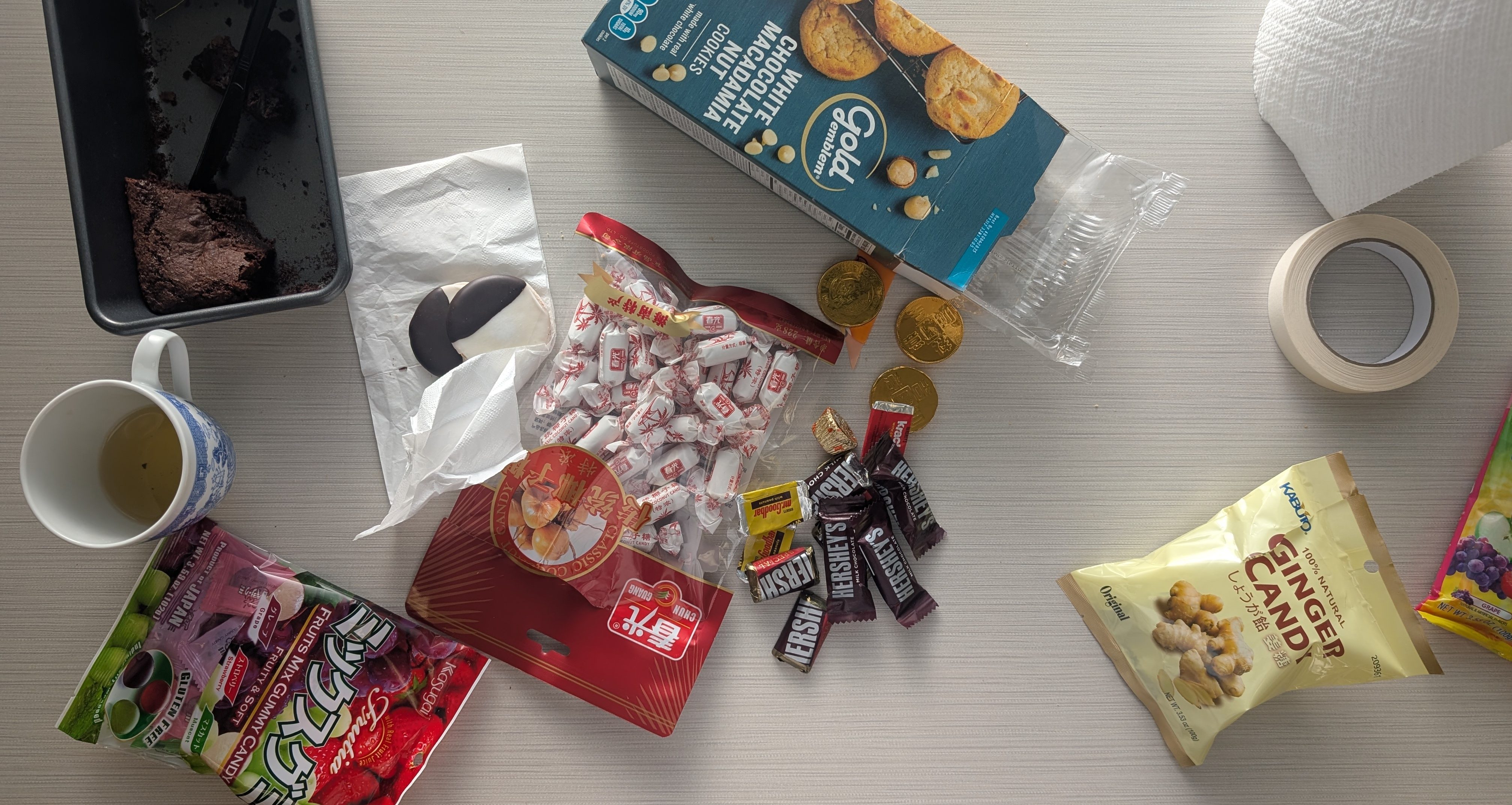 PXL_20250314_221910319.MP GenAI Alt text: Various snacks including half-eaten chocolate cake, black and white cookies, wrapped candies, Hershey's miniature chocolates, and a cup with tea on a light-colored table